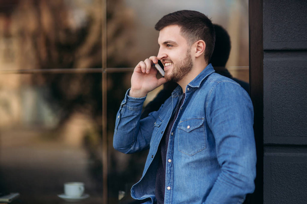 Knappe man in een overhemd denim maakt gebruik van een telefoon staan op straat - Foto, afbeelding
