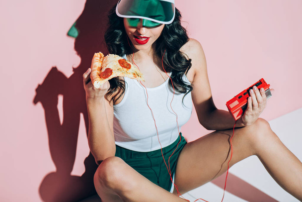 stylish woman in cap with retro music player holding piece of pizza on pink backdrop - Foto, Bild
