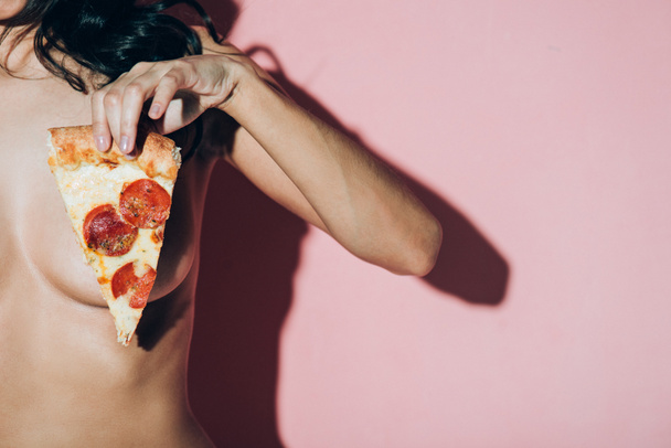 částečný pohled topless ženy držící kousek pizzy na růžové pozadí - Fotografie, Obrázek