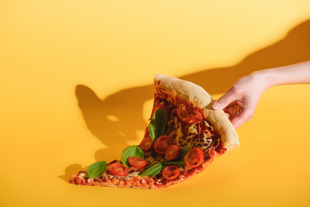 Teilansicht einer Frau, die ein Stück Pizza mit Kirschtomaten in der Hand auf gelbem Hintergrund hält - Foto, Bild