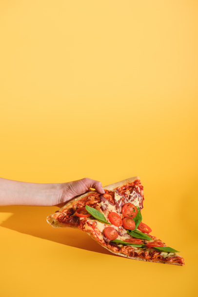 partial view of woman holding piece of pizza with cherry tomatoes in hand on yellow background - 写真・画像