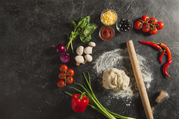 flat lay with rolling pin, raw dough and fresh ingredients for pizza on dark surface - Фото, зображення
