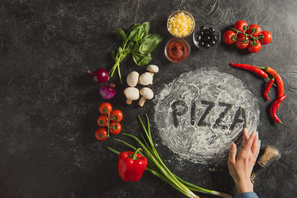 partial view of female hand, fresh ingredients and pizza lettering made of flour on dark surface - Photo, Image
