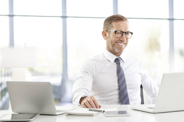 Uomo d'affari di successo indossando camicia mentre seduto alla scrivania dell'ufficio e lavorando su computer portatili
.  - Foto, immagini