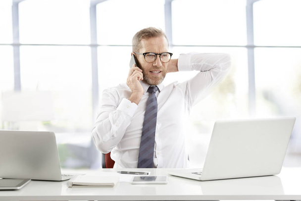 Конфліктний бізнесмен середнього віку в сорочці і краватці і робить дзвінок, сидячи перед ноутбуком і працюючи в офісі
.  - Фото, зображення