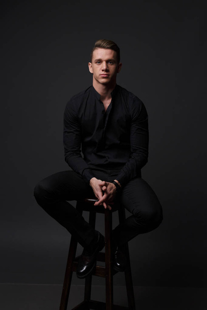 a guy in a black shirt and black pants sits on a dark chair, on a gray background - Fotoğraf, Görsel