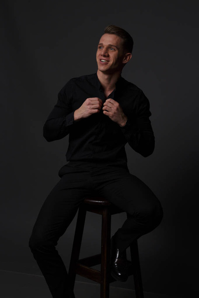 a guy in a black shirt and black pants sits on a dark chair, on a gray background - Фото, зображення