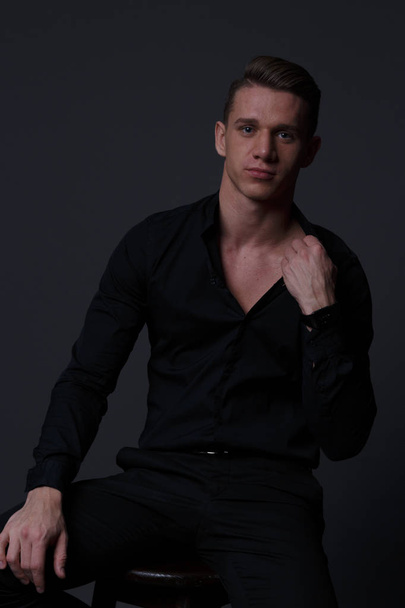 a guy in a black shirt and black pants sits on a dark chair, on a gray background - Foto, Bild