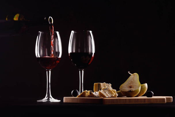 wine glass and bottle, cheese with mold, pear and grapes on black background - Photo, Image