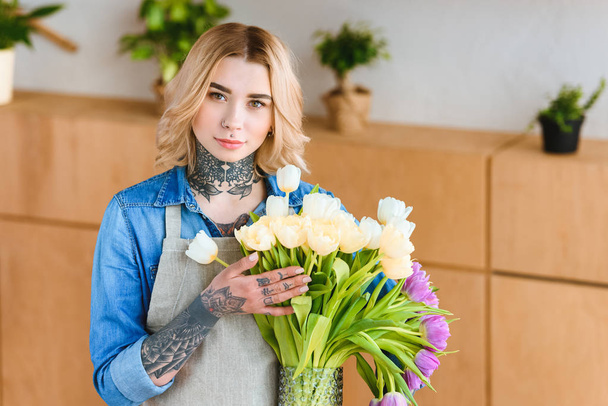young female florist in apron holding beautiful tulips in vase and looking at camera - Photo, Image