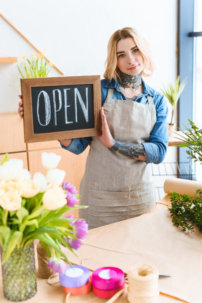 beautiful young florist holding open sign and looking at camera in flower shop - Foto, imagen