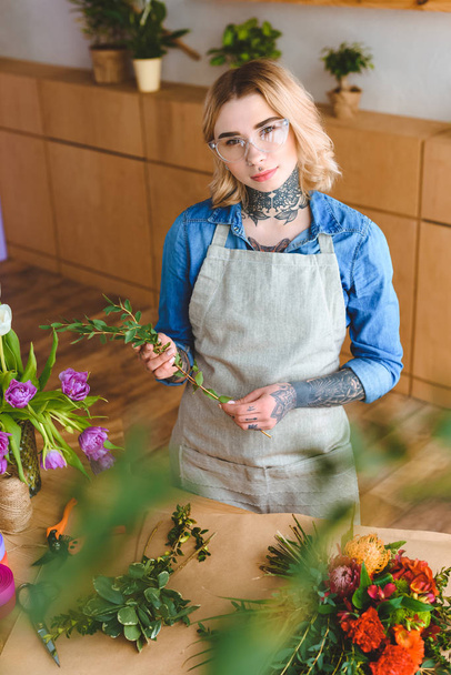 beautiful young florist in apron and eyeglasses arranging flowers and looking at camera - 写真・画像