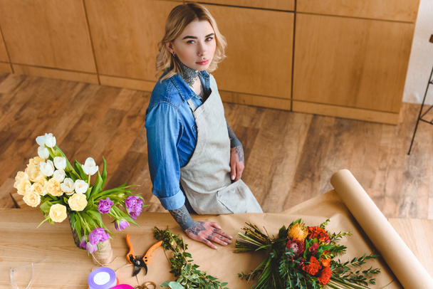 high angle view of beautiful young florist in apron standing at workplace and looking at camera - Photo, Image
