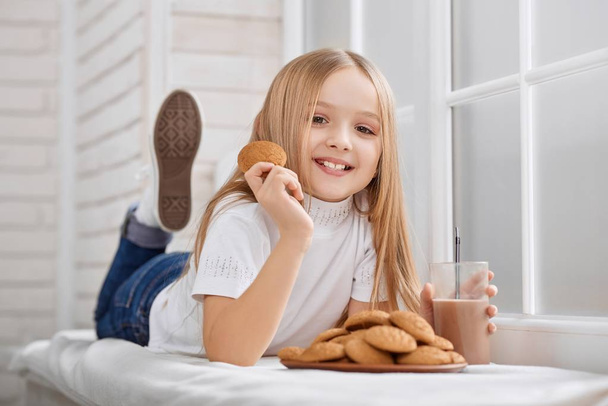 Little girl lies on window sill with cookies and chocolate milk. - Фото, изображение