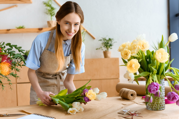 beautiful young florist wrapping tulips in craft paper and smiling at camera - Photo, Image