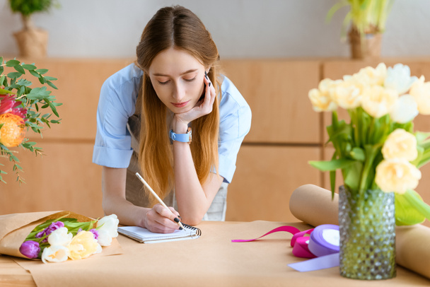 beautiful young florist taking notes in notebook at workplace  - Photo, image