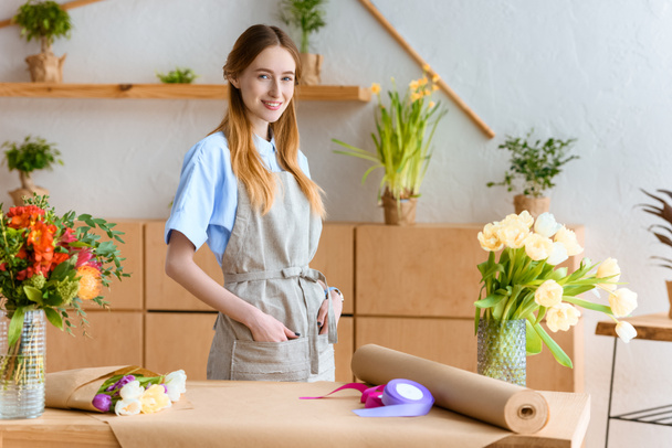 young florist in apron standing with hands in pockets and smiling at camera in flower shop - Photo, image