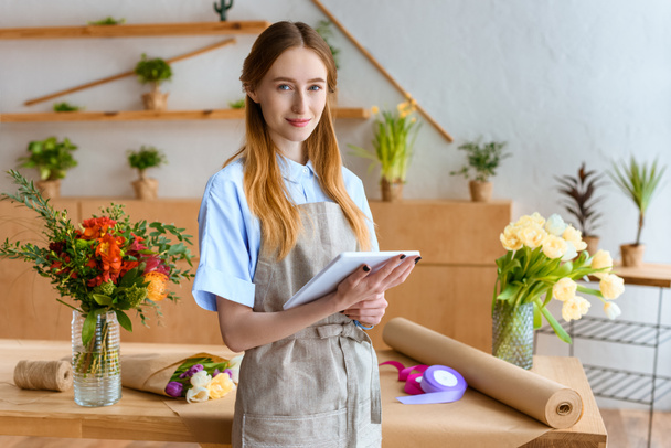 beautiful young florist holding digital tablet and smiling at camera in flower shop - Fotoğraf, Görsel