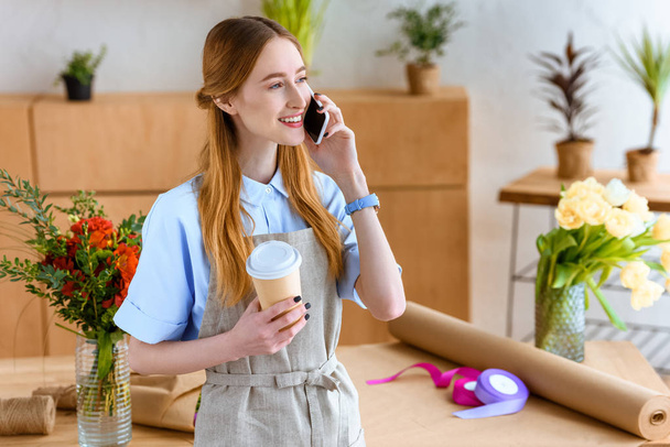Улыбающийся молодой флорист держит кофе, чтобы пойти и поговорить со смартфоном
 - Фото, изображение