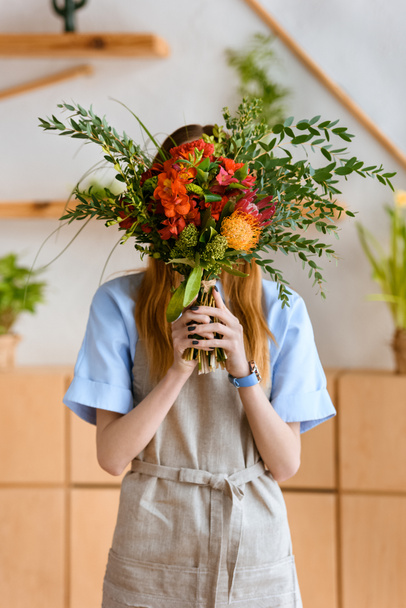 young florist in apron hiding face behind beautiful flower bouquet  - Foto, afbeelding