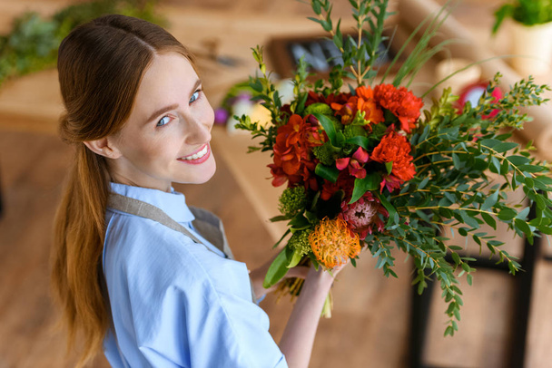 high angle view of beautiful young florist holding bouquet of flowers and smiling at camera - 写真・画像