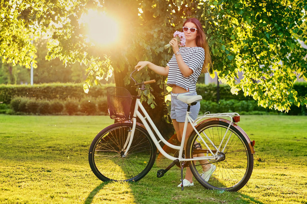 Beautiful brunette stands on the green lawn with bicycle in park - Foto, imagen