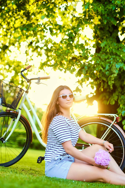 Hermosa morena se sienta en el césped verde con bicicleta y sostiene
 - Foto, Imagen