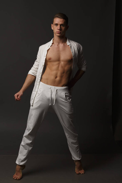 a guy with a naked torso, in white pants, and in a white shirt, stands on a gray background - Фото, зображення