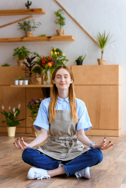 smiling young florist in apron sitting in lotus position on floor in flower shop - Foto, Imagen