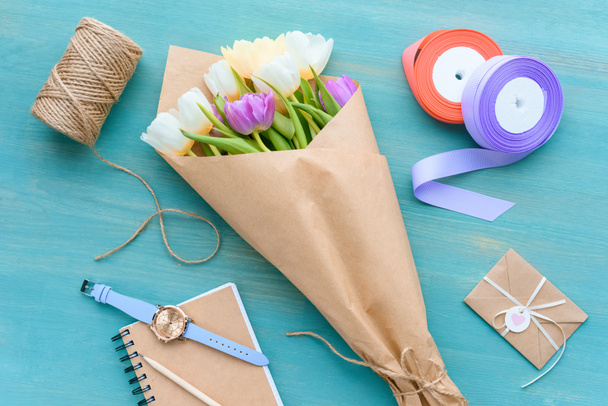top view of beautiful tulips in craft paper, rope, ribbons, envelope and notebook with wristwatch - Fotografie, Obrázek