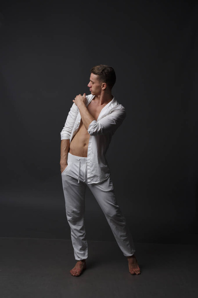 a guy with a naked torso, in white pants, and in a white shirt, stands on a gray background - Φωτογραφία, εικόνα