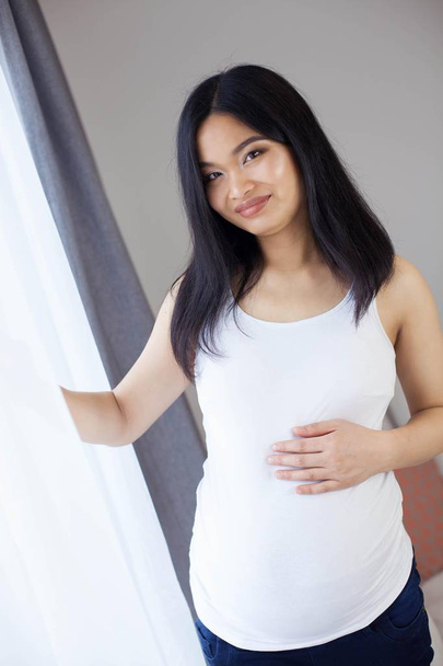 Piękna młoda Azjatka w ciąży kobieta stojąc przy oknie i dotykając jej brzuch. Ciąży. Zdrowia. Macierzyństwo. Przytulne portret kobiety w ciąży. Cozy. - Zdjęcie, obraz