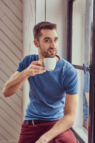 Young caucasian student drinks morning coffee sitting on a windo - Foto, Imagem