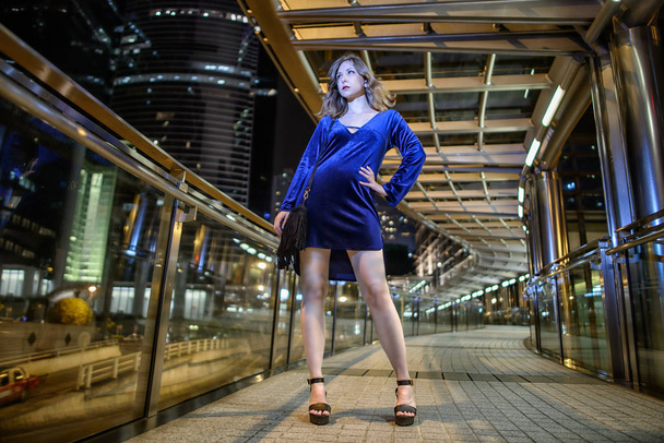 Beautiful slender young girl model with brown hair and long legs in a blue short velvet dress, with a black little bag with fringe poses in soffit light. Fashion style photo shoot in night big city Hong Kong - Photo, Image