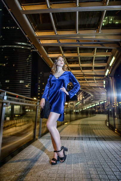 軒天井の光のフリンジ ポーズと黒の小さな袋で茶色の髪と青の短いベルベット ドレスの長い脚と美しいスレンダー少女モデル。夜大都市香港のファッション スタイル写真撮影 - 写真・画像