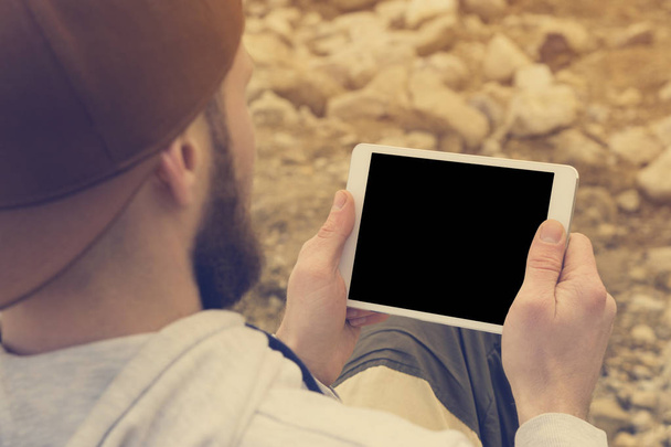 Κοντινό πλάνο του μια ορδή σε ένα καφέ καπάκι στον ανοιχτό αέρα κρατά ένα λευκό tablet pc στα χέρια του. Ένας γενειοφόρος άνδρας κοιτάζει το tablet. OTS θέα από πίσω από τον ώμο - Φωτογραφία, εικόνα