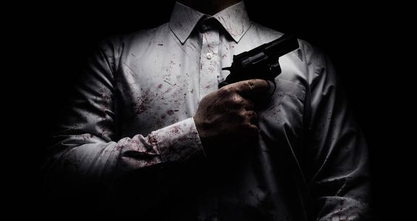Horror scary photo of a killer in white shirt with blood splatter and posing with black gun on dark background. - Photo, Image