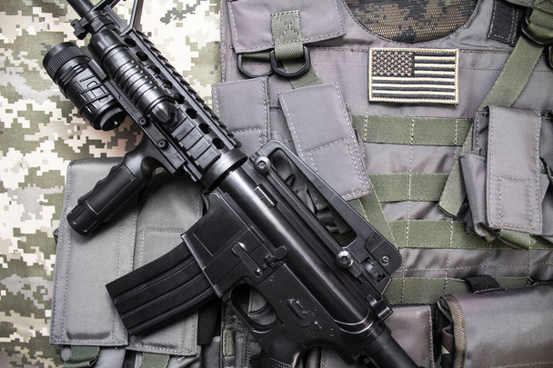 Foto vista superior de um colete à prova de balas militar com crachá de bandeira americana e rifle colocado em um fundo de pano de camuflagem
, - Foto, Imagem