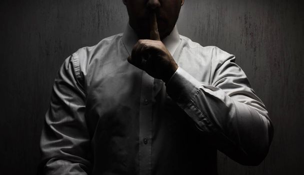 Foto de um homem de terror assustador em camisa branca em pé sob a luz superior no fundo preto mostrando sinal de mão silêncio
. - Foto, Imagem