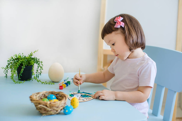 niño pinta huevos en huevos de madera sentados a la mesa en una habitación blanca - Foto, imagen