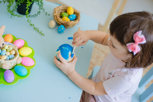 niño pinta huevos en huevos de madera sentados a la mesa en una habitación blanca - Foto, Imagen