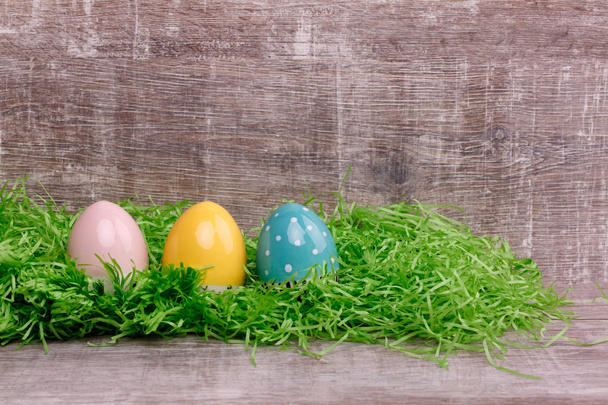 Three bright colorful eggs on green grass in front of wooden background. Easter greeting card.  - Foto, immagini