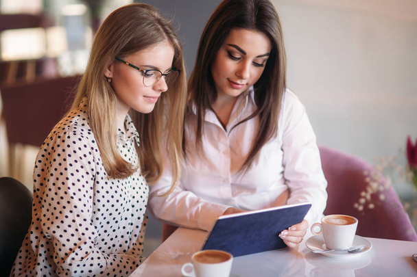 Deux filles utilisent une tablette tout en étant assis dans un café et en buvant du café
 - Photo, image