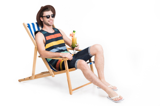 schöner Mann mit Sonnenbrille und Hut entspannt im Strandkorb mit Cocktail, isoliert auf weiß - Foto, Bild
