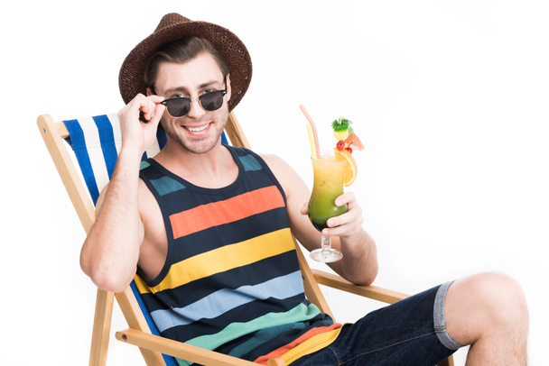 güneş gözlüğü ve şapka kokteyl ile plaj sandalyesi üzerinde rahatlatıcı adam gülümsüyor, üzerinde beyaz izole - Fotoğraf, Görsel