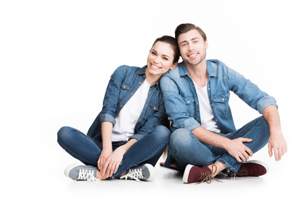 cheerful couple sitting in jeans and looking at camera, isolated on white - Фото, изображение