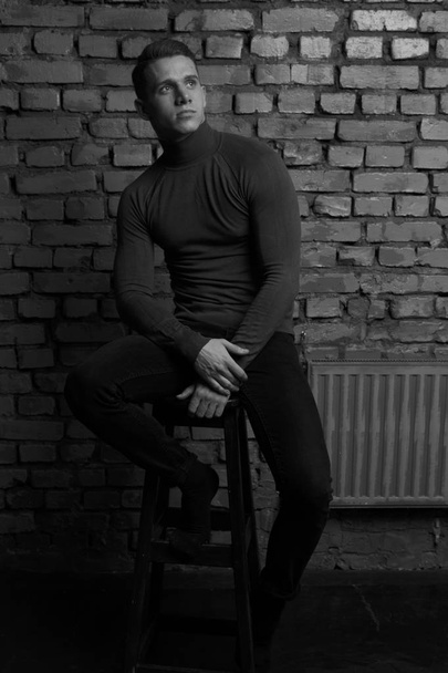 white young guy sitting on a chair near a gray brick wall, black-and-white photo - Fotografie, Obrázek