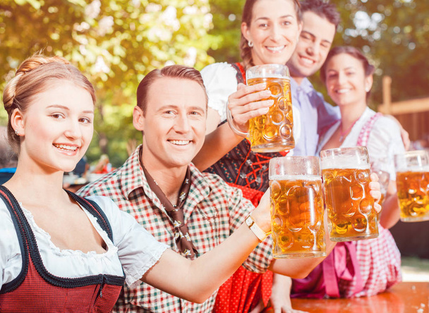Amigos vistiendo Tracht, Dirndl y Lederhosen y bebiendo cerveza fresca en In Beer garden, Bavaria, Alemania
. - Foto, Imagen