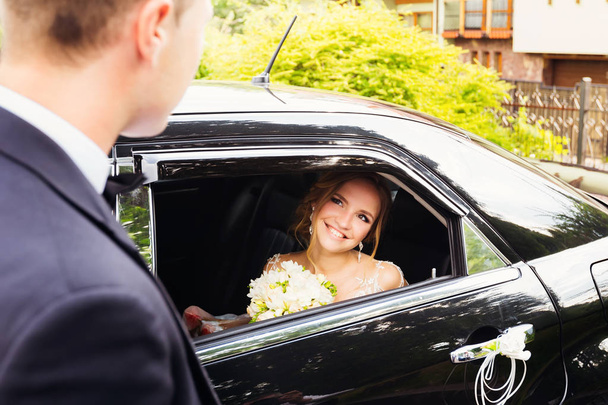 Νύφη κάθεται σε ένα μαύρο αυτοκίνητο, και κοιτάζοντας τον μνηστήρα της μέσα από ένα παράθυρο - Φωτογραφία, εικόνα