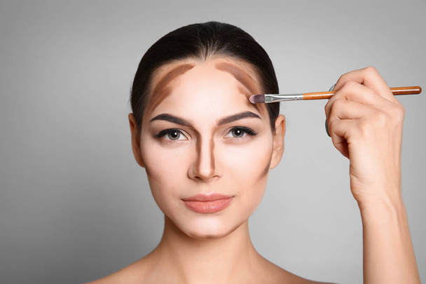 Young woman applying contouring lines on grey background. Professional makeup products - Photo, Image